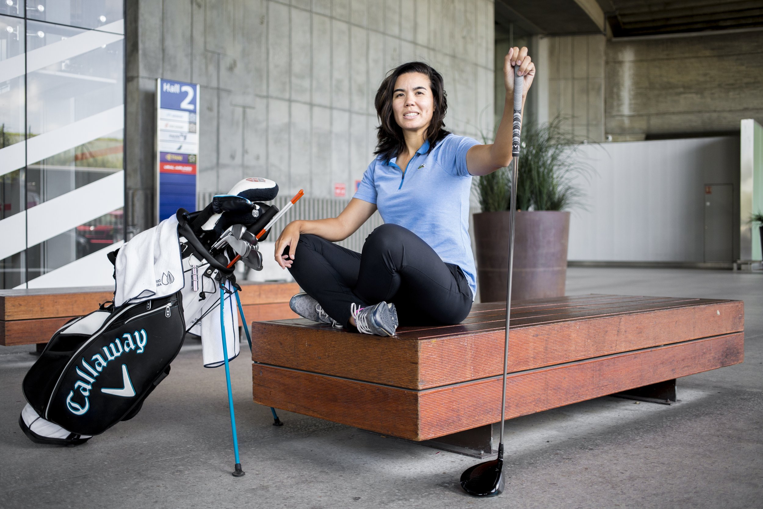 A woman in profile seated on an airport bench, wearing a blue polo shirt and black pants, holding a golf club in one hand and a golf club in the other, with a golf bag at her side.