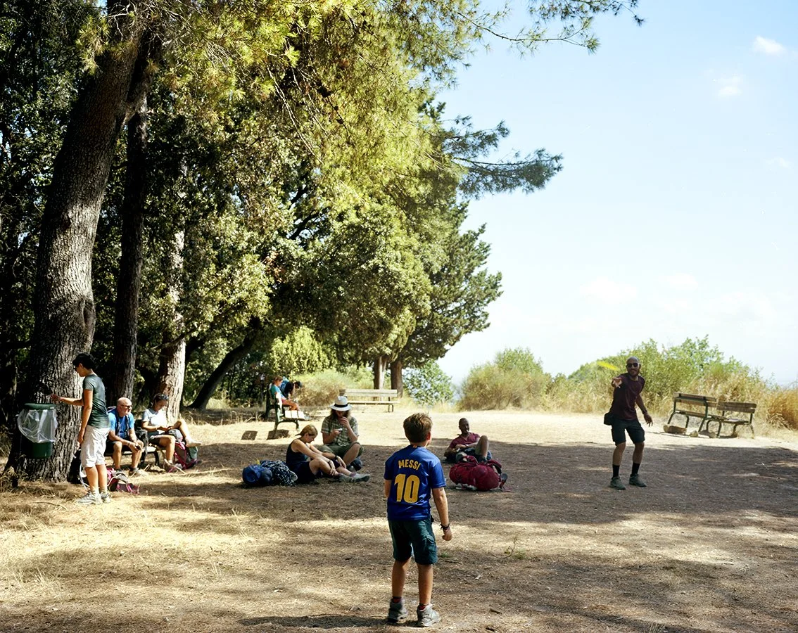 Group of people, including children, sitting and standing under trees in a park, with a guide or monitor talking or explaining something.