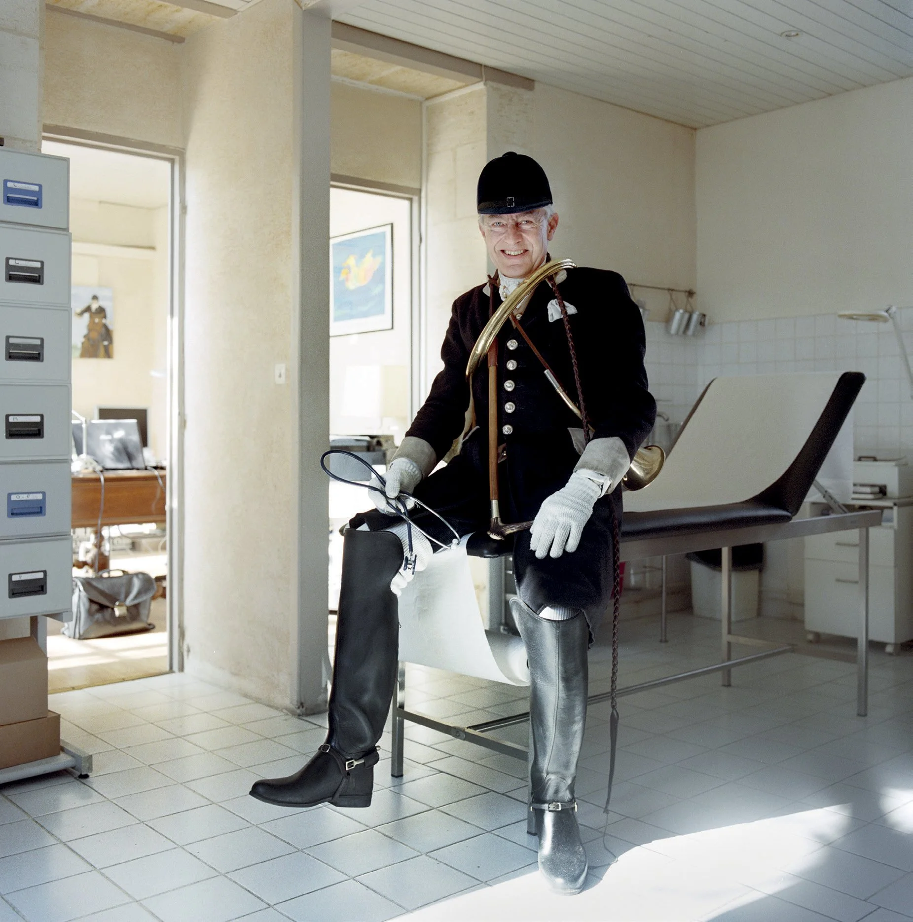 A man in historical costume with a long black robe, black cap and silver-effect leather boots sits at a medical examination table in a white room. He carries a horn and hunting horn, holding a pair of glasses, da