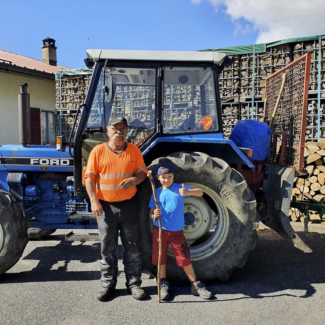 An adult man and a young boy pose in front of a blue tractor, with a pile of wood in the background. The man is wearing an orange t-shirt with reflective stripes, while the boy is wearing a blue t-shirt and red pants. The scene takes place in daylight