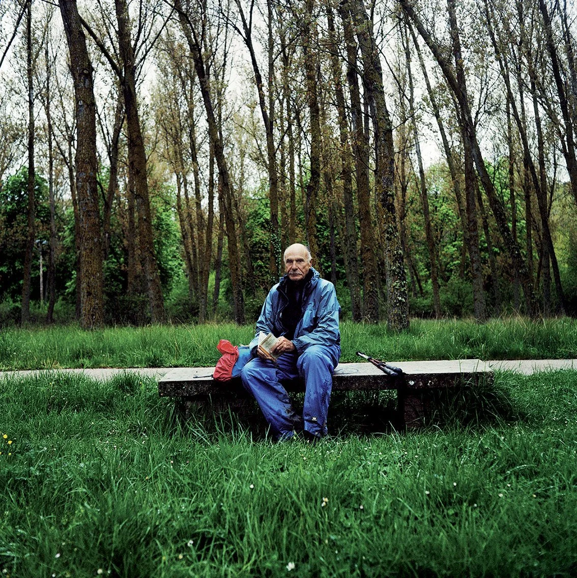 An elderly man sitting on a wooden bench in a forest, holding a book, with a red tarpaulin, umbrella and bag beside him.