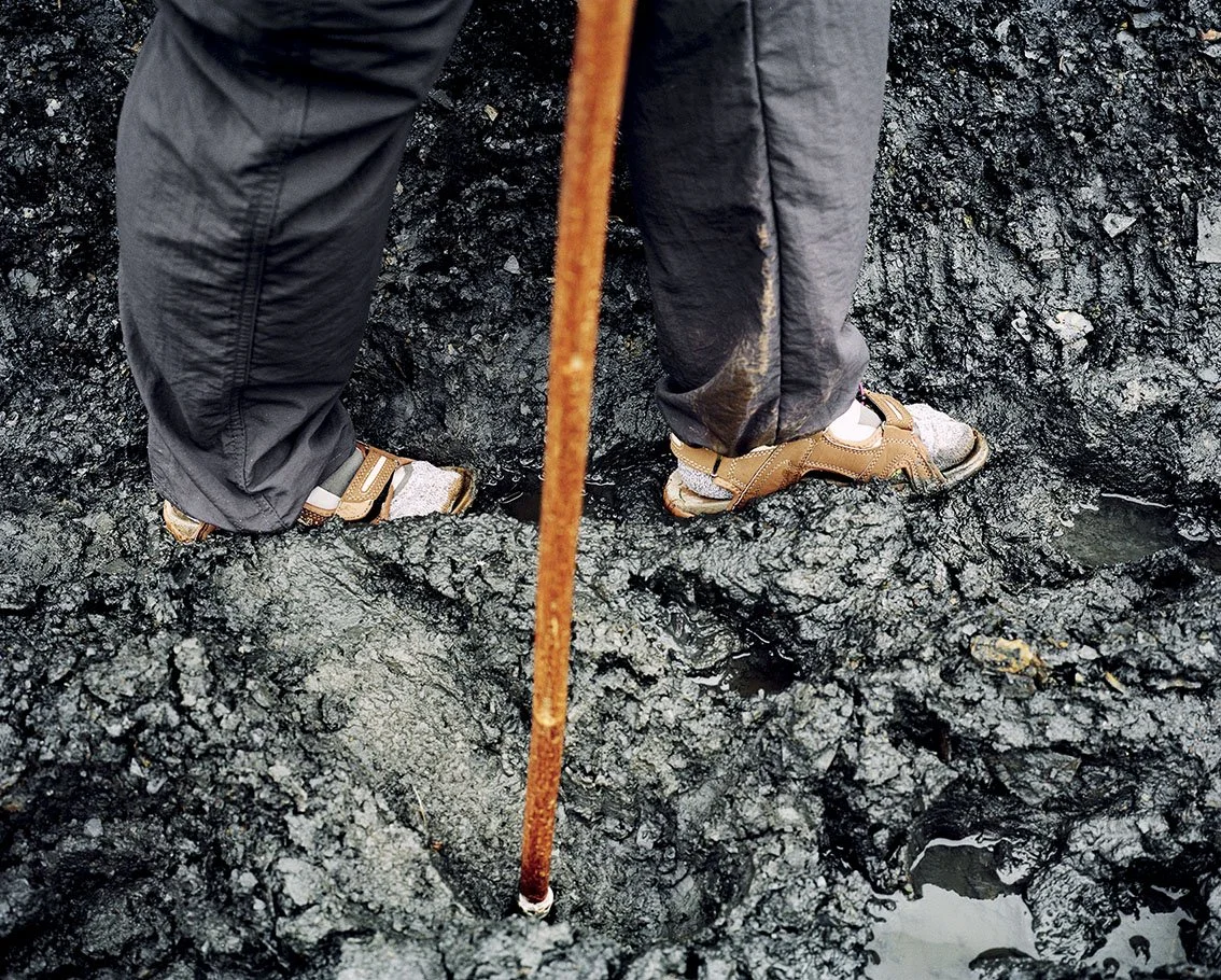 A person wearing hiking boots and protective clothing, walking on solid black volcanic lava, with a cane or wooden stick at his side.