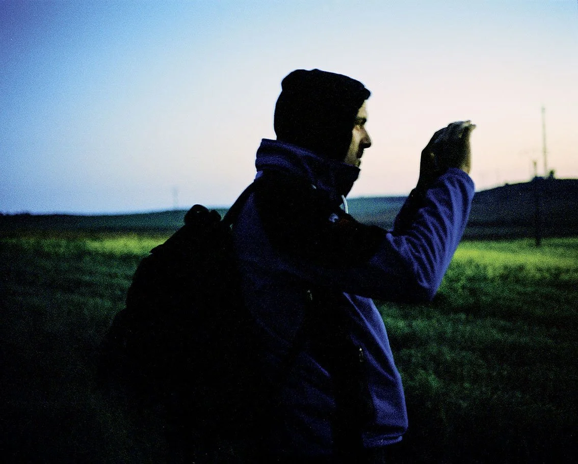 Young man in profile wearing a jacket, raising his hand in a field at dusk.