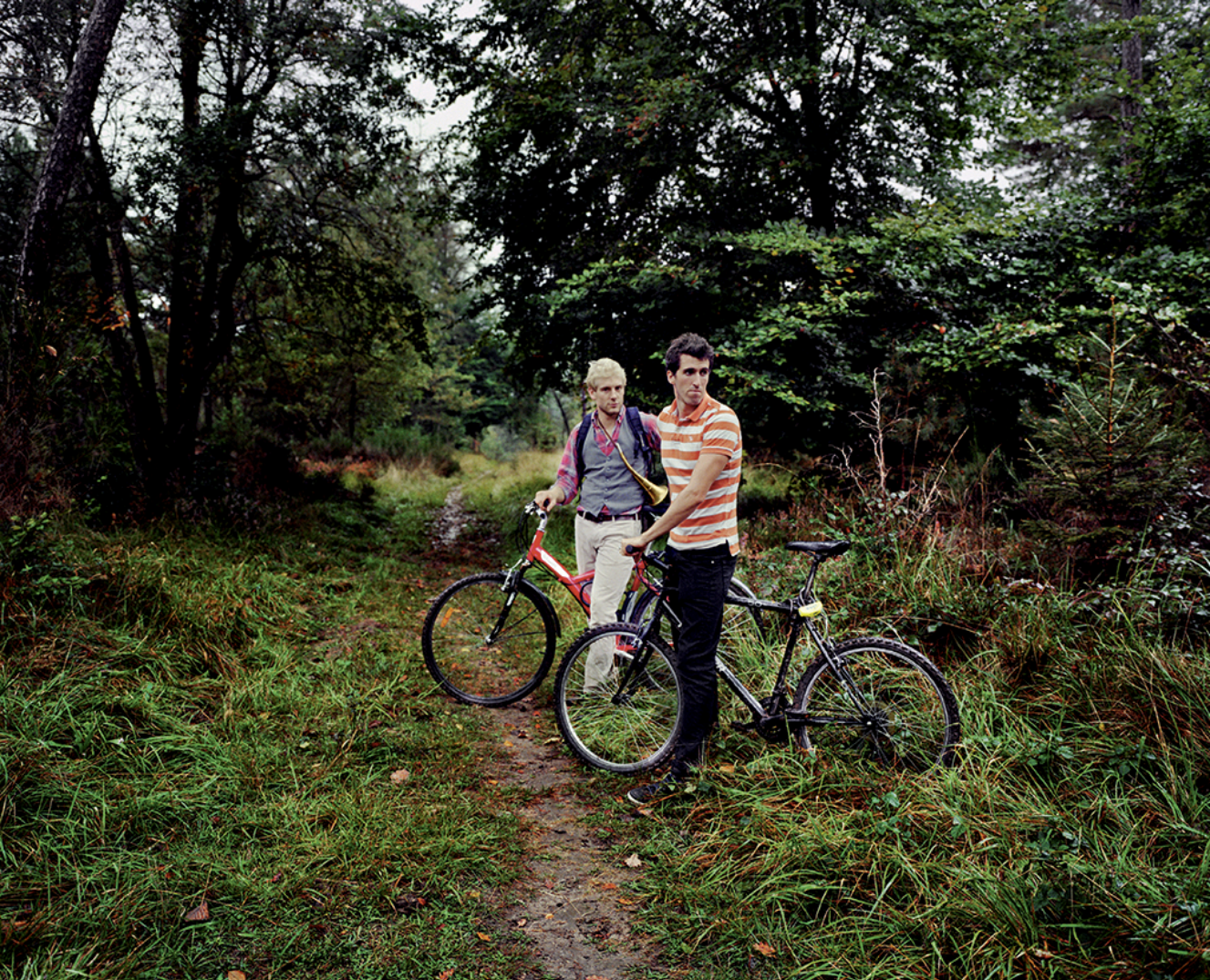 Deux jeunes hommes avec des vélos dans une forêt dense, un chemin de terre, arbres et végétation luxuriante.