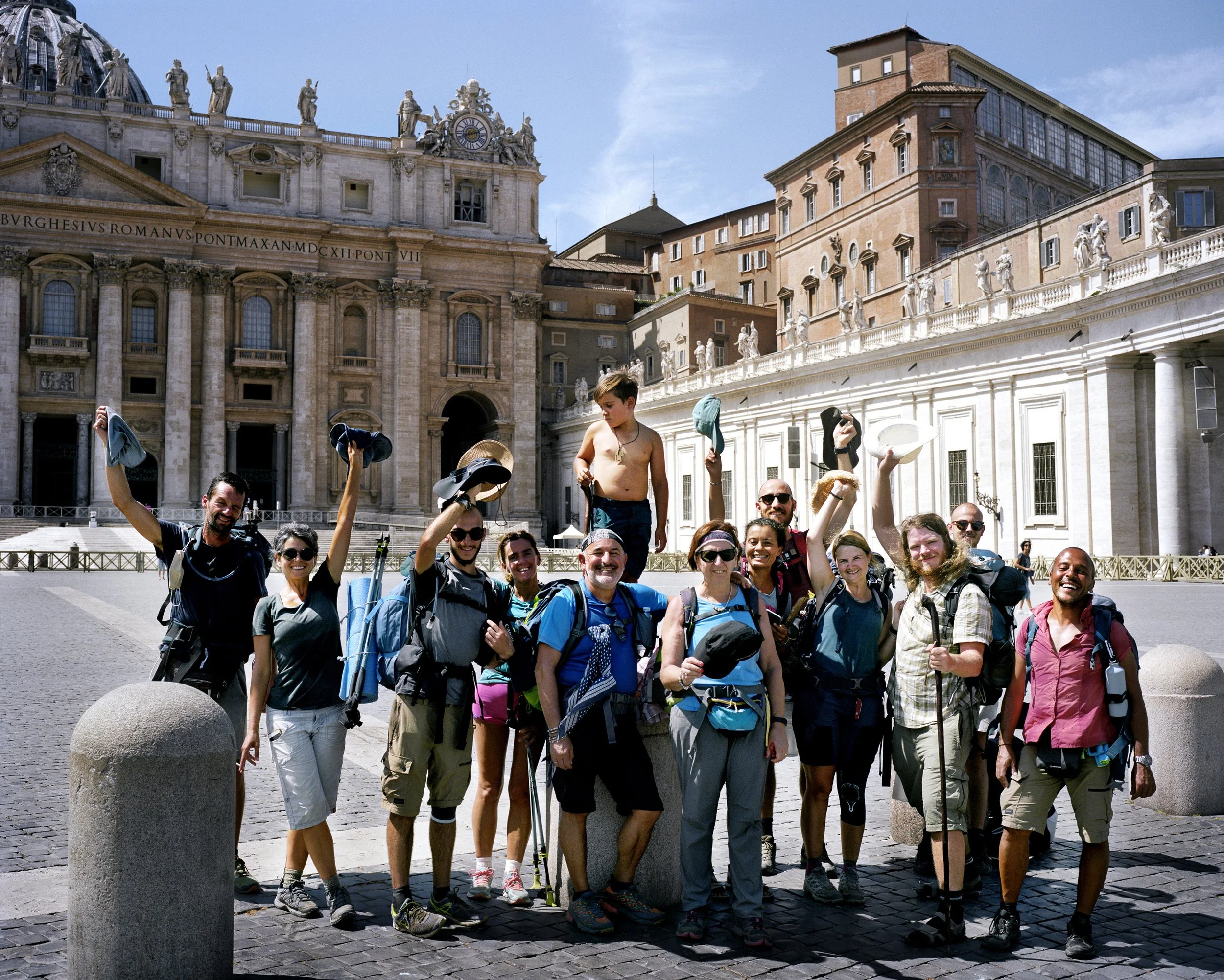 A group of hikers pose for a photo in front of a historic monument in Rome, with a church adorned with statues and a stone facade in the background, under a blue sky.