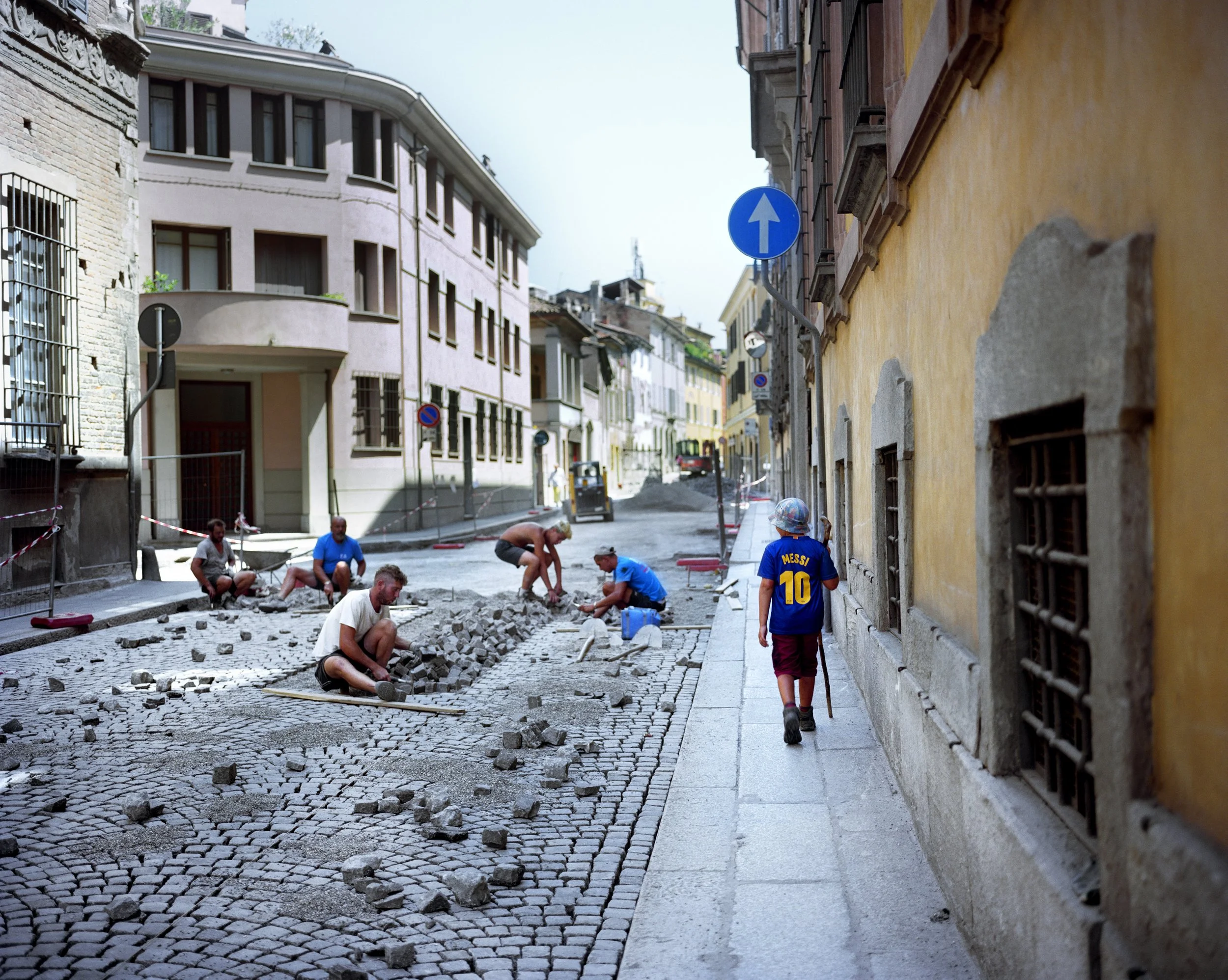 Cobblestone streets under renovation with workers on the pavement, a teenage girl wearing a Messi soccer jersey, and a blue directional sign.