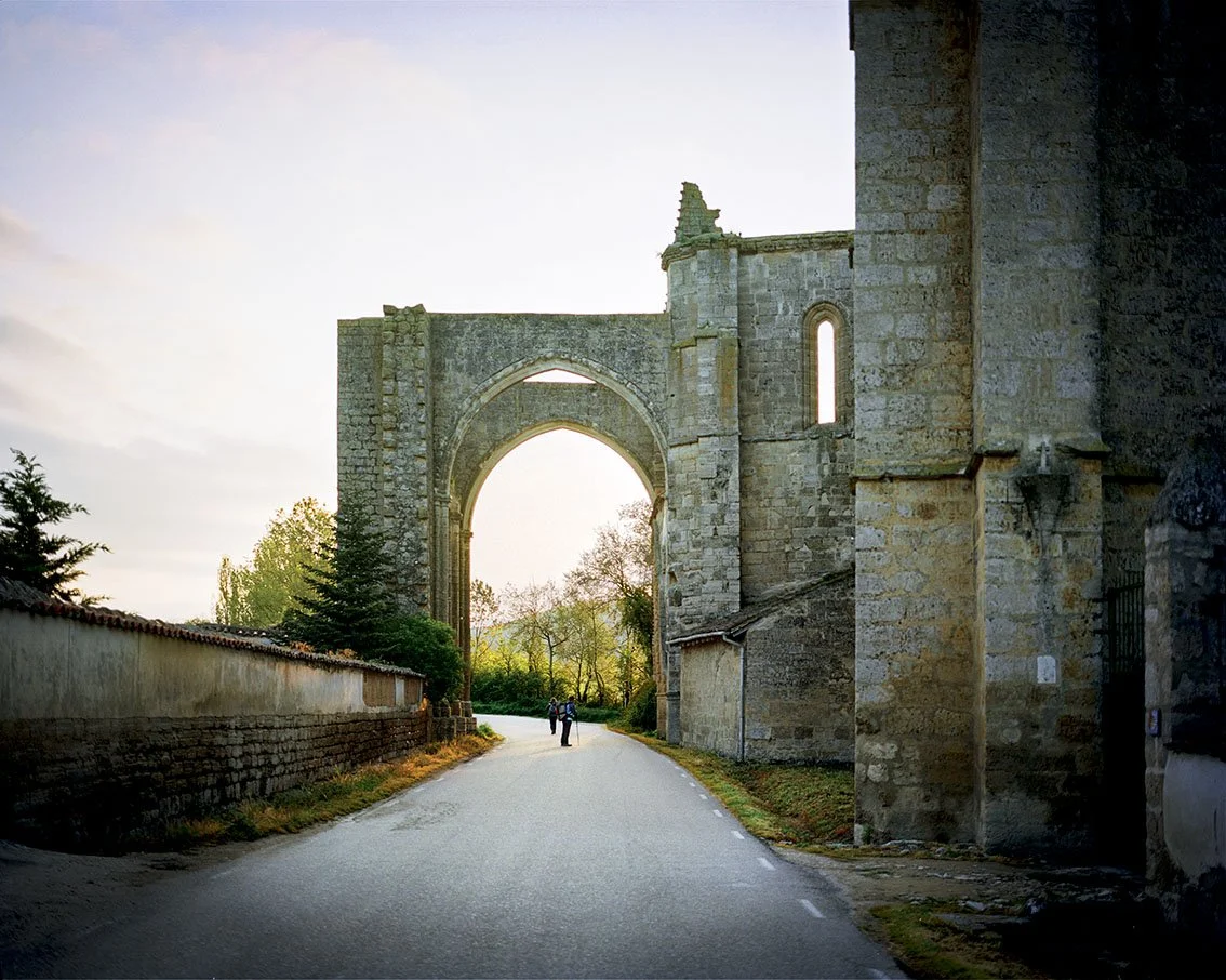 Ruins of ancient stone architecture with a large arch and narrow windows, seen from a road with two people walking, at dusk.
