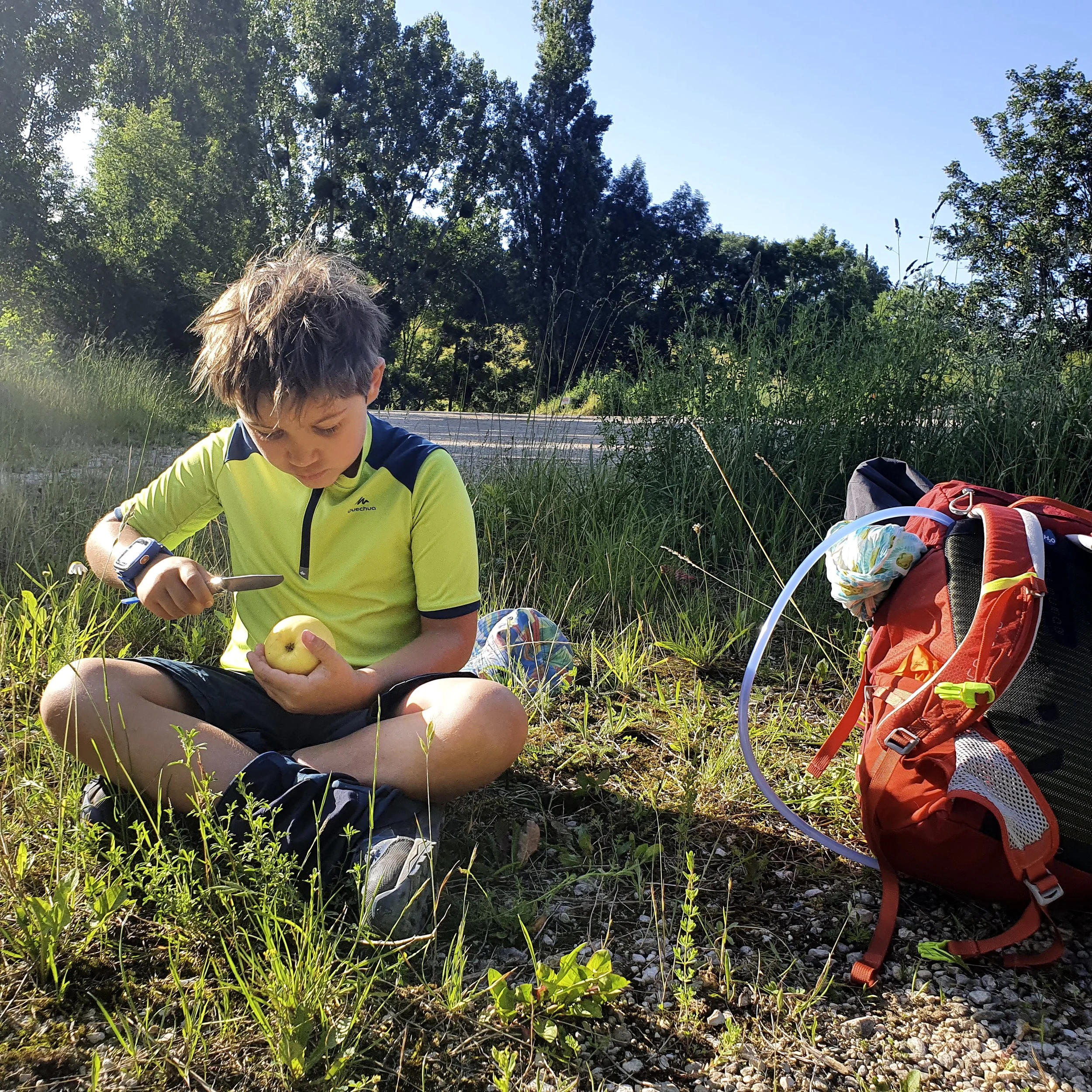 Young boy sitting on the grass, using a knife to cut an apple, gadget bracelet on his wrist, red backpack at his side, outdoors with trees in the background.