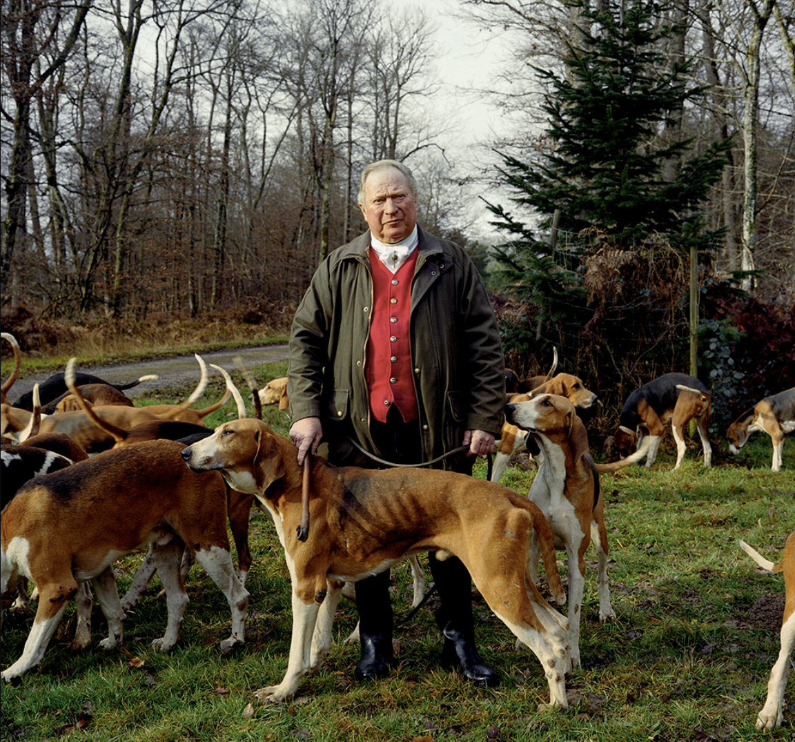 Un homme avec un manteau vert, tenant la laisse d'une dogue allemand, entouré de plusieurs autres chiens dans une forêt en automne.