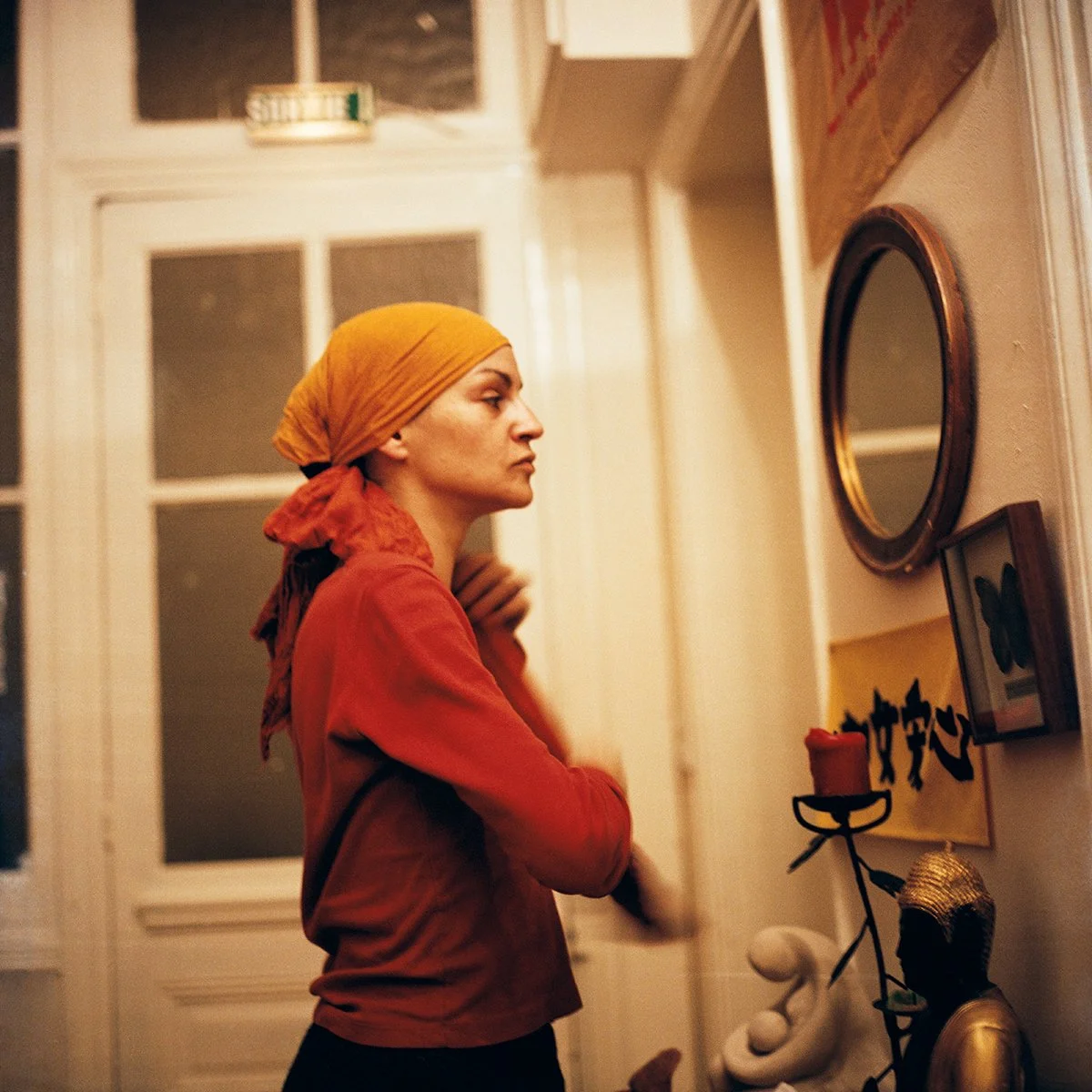 Femme avec un foulard orange regardant un miroir, décor intérieur avec bougies et œuvres d'art.