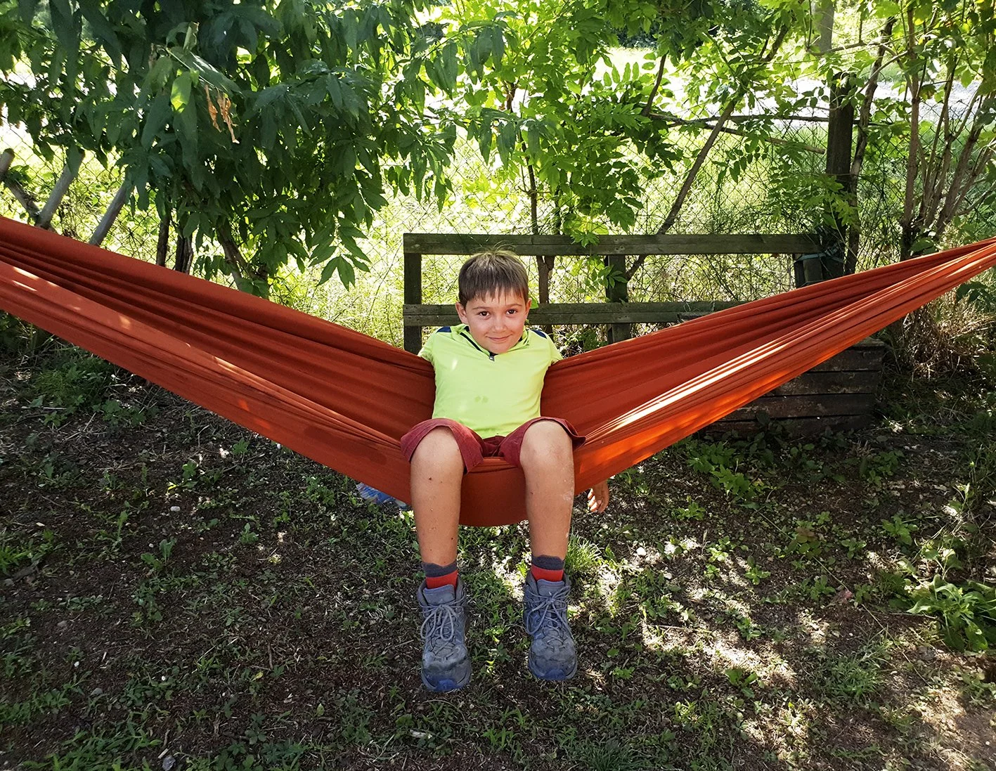 A boy sitting in an orange hammock suspended between two green trees in a garden.