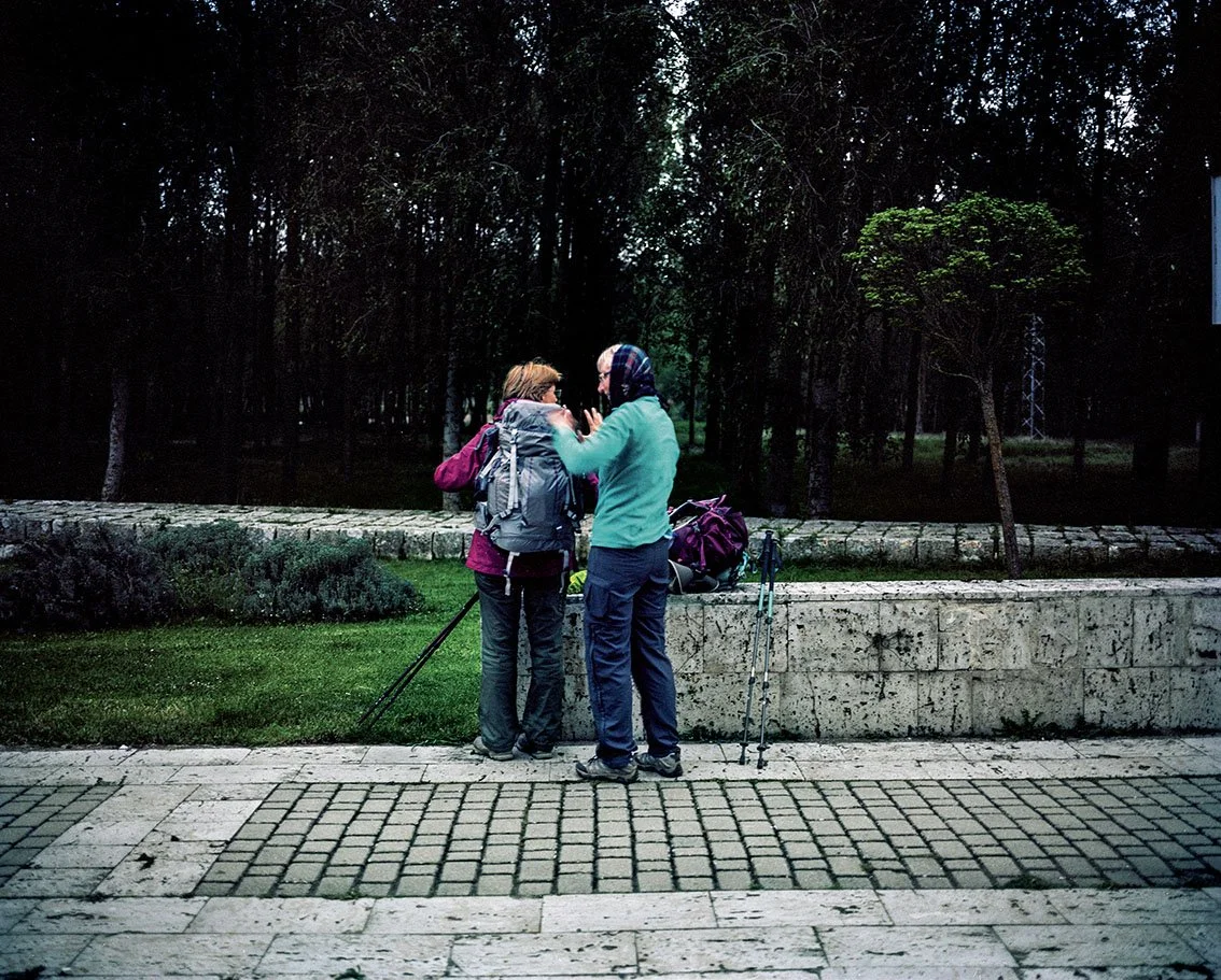 Two women equipped with backpacks and walking sticks are chatting in a park, with trees in the background.