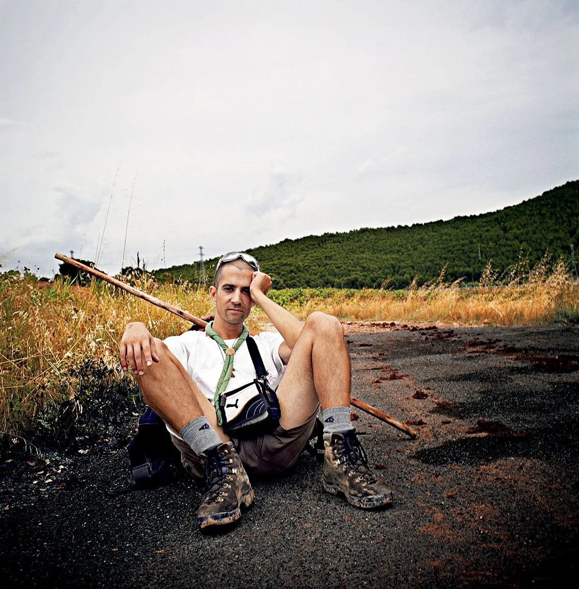 A man sitting on the ground in a rural area, wearing hiking clothes, with a stick used for walking, a pair of glasses on his head and a camera hanging around his neck, in a landscape of fields and hills.