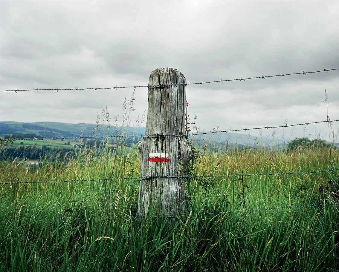 Wooden post with painted red line, separating a field of green grass with a landscape in the background and a cloudy sky by a barbed wire fence.