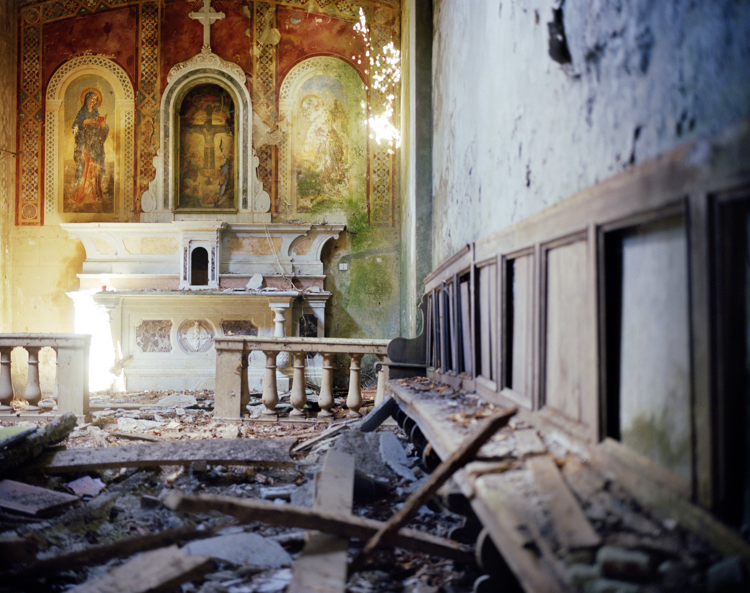 Interior of an abandoned church with a damaged altar, walls partially covered with ancient frescoes, a debris-strewn floor and a row of damaged wooden pews.