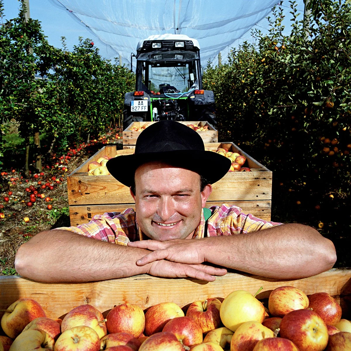A smiling man wearing a black hat, posing on an apple farm, with crates full of apples, a tractor in the background, in a rural setting in broad daylight.