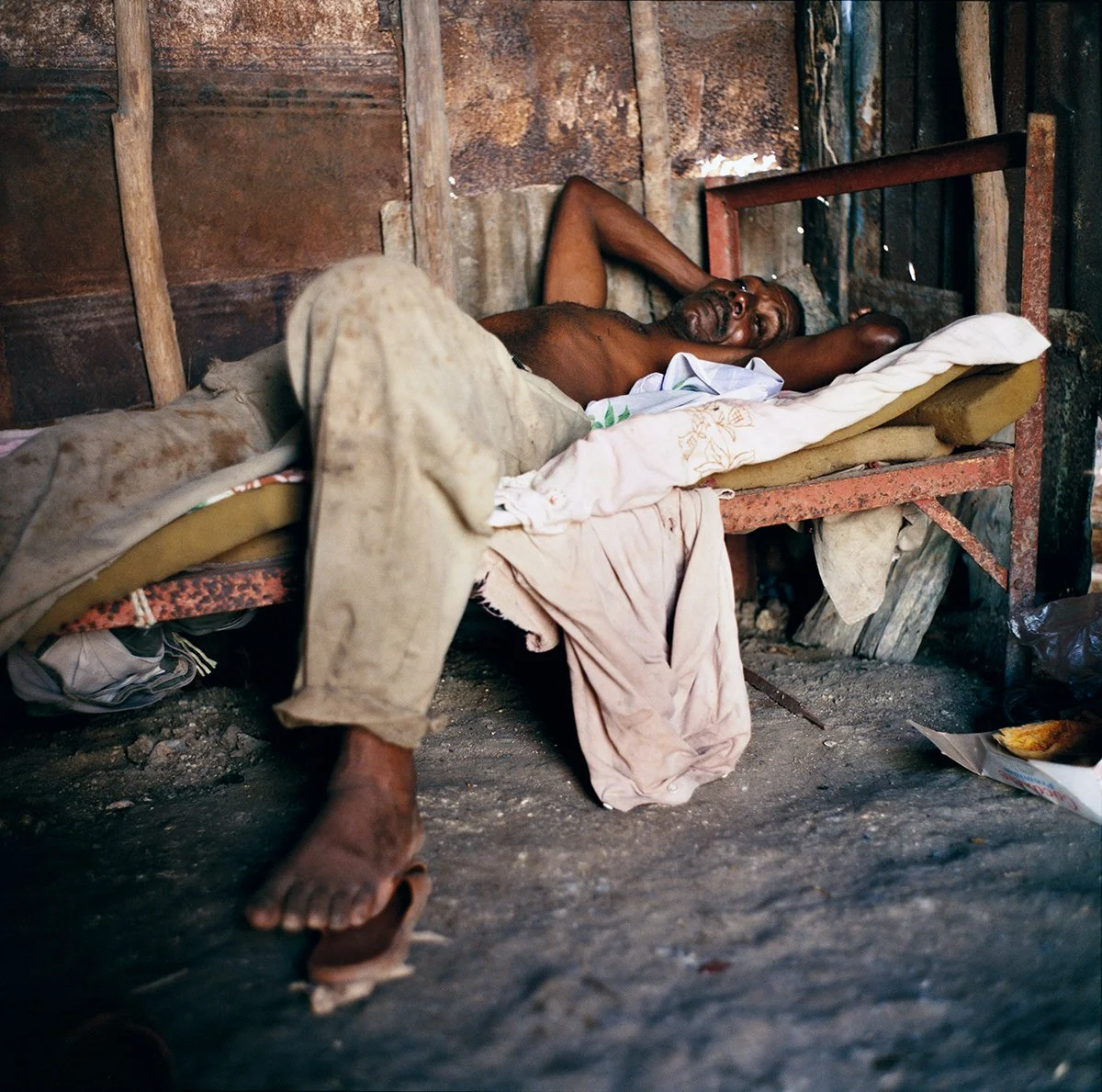 A man lies on a makeshift bed in a rudimentary room with wooden walls. He appears to be sleeping or resting, dressed in worn clothing.