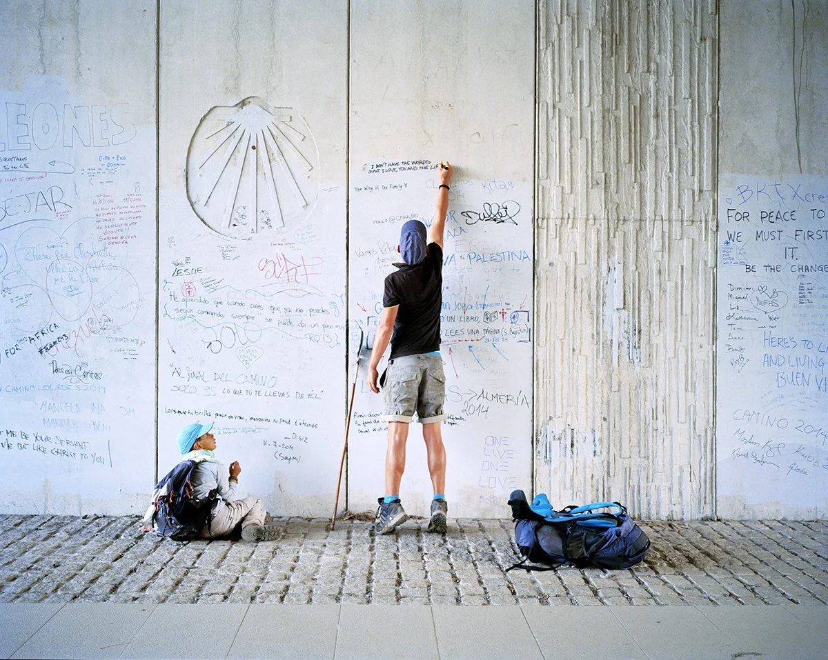 Two young people, one sitting on the ground and the other standing, write on a white wall graffitied with messages and drawings. The seated one wears a blue hat, backpack and light clothes, while the standing one, in shorts and black T-shirt, writes s