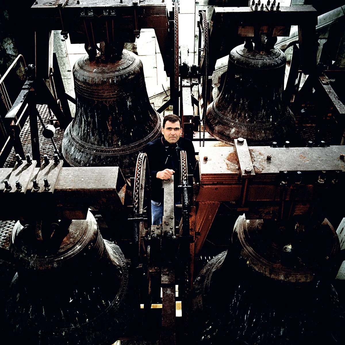 A man standing in a tower with large metal bells, probably in a clock tower or giant bell.