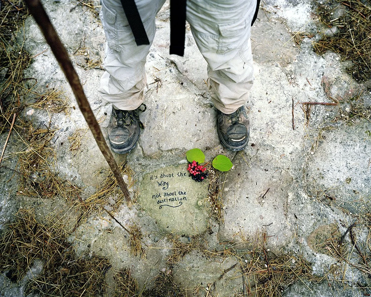 Man in hiking boots and beige pants, seen from above, standing in front of a stone engraved with a quotation and red flowers. The text reads: "It's about the way not about the destination".