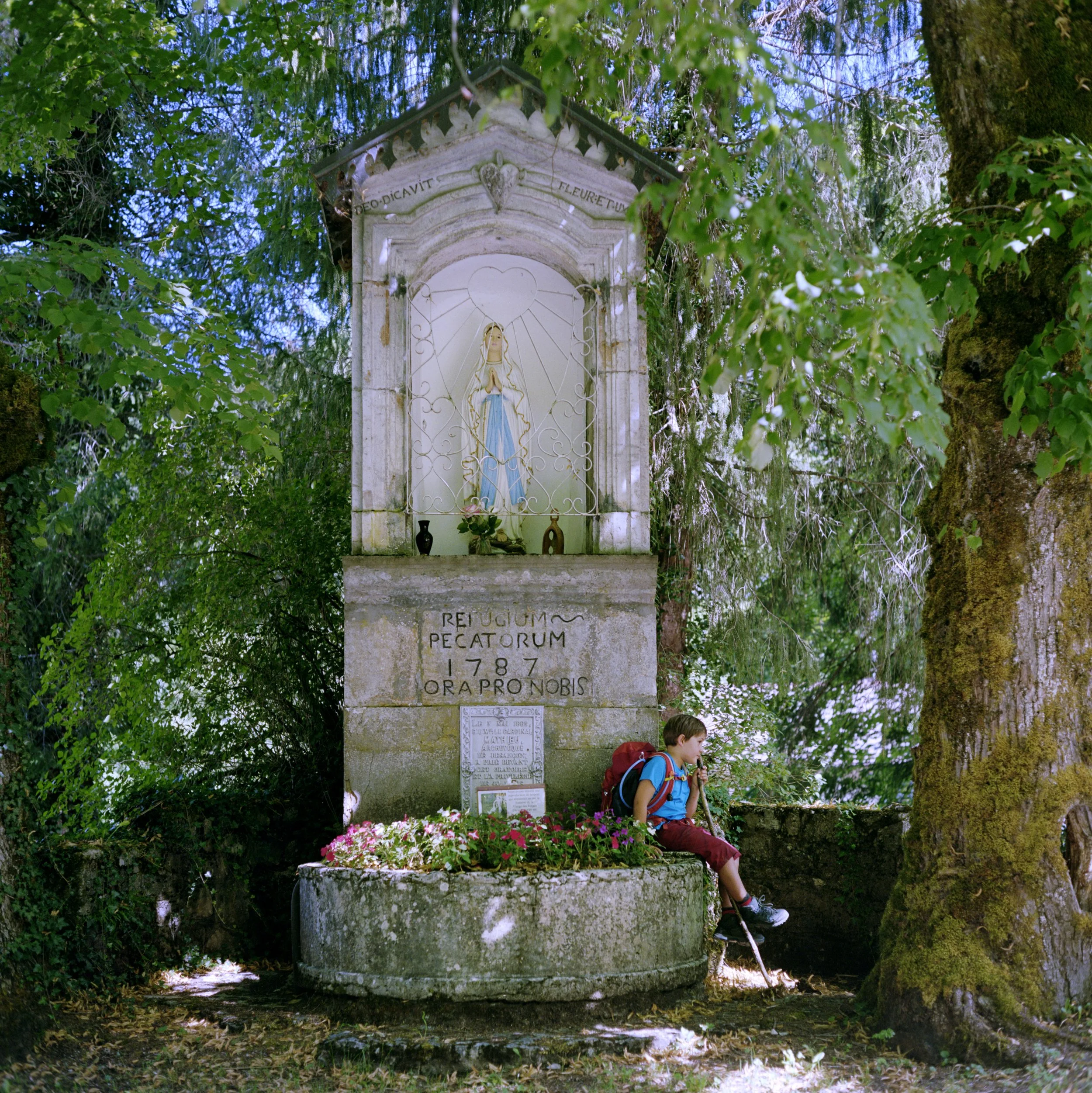 Young boy with backpack sitting next to a religious monument dedicated to Mary, surrounded by trees and flowers in a forest setting.