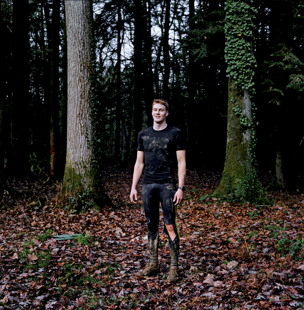 Jeune homme debout dans une forêt, vêtu de vêtements sales et endommagés, entouré d'arbres et de feuilles mortes.