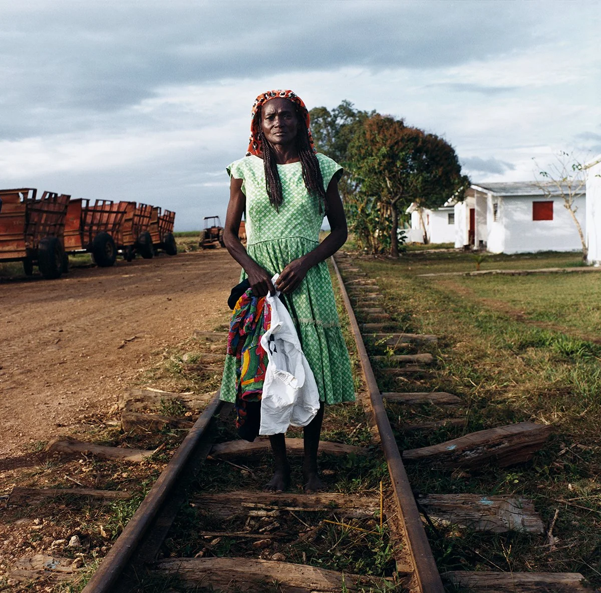 Woman standing on railroad tracks, holding clothes in her hands, with a rural landscape in the background.