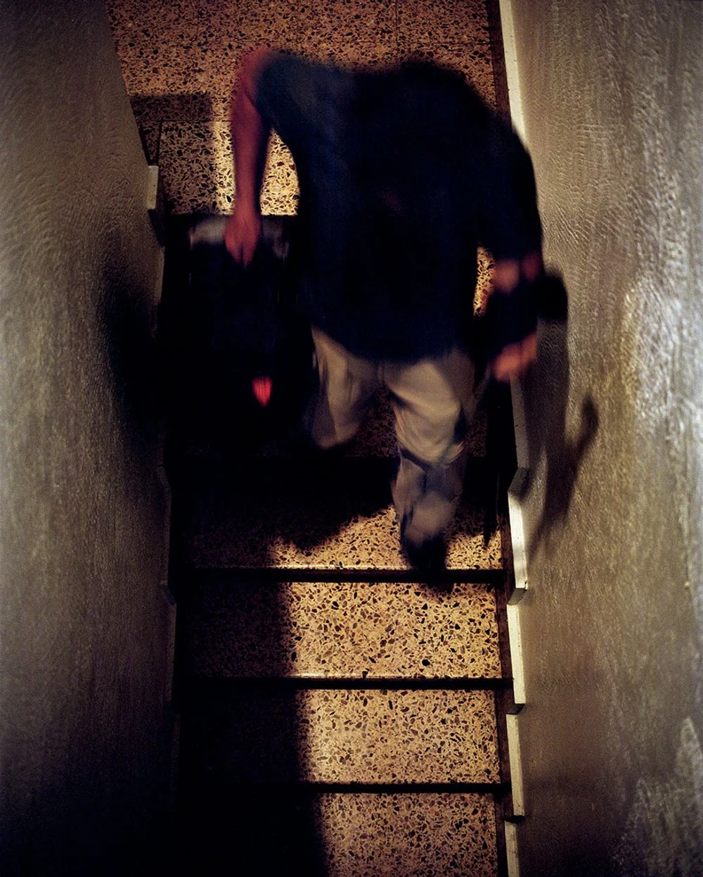 A person descends a narrow, dark staircase, carrying a black handbag with a red heart motif on it.