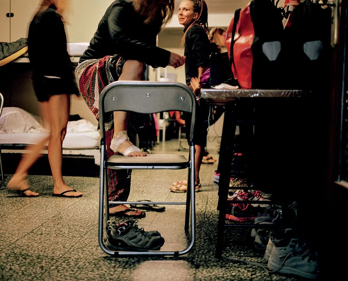 Young girls in beachwear or briefs, getting ready in a room, some wearing shoes or sandals, with backpacks on a table.