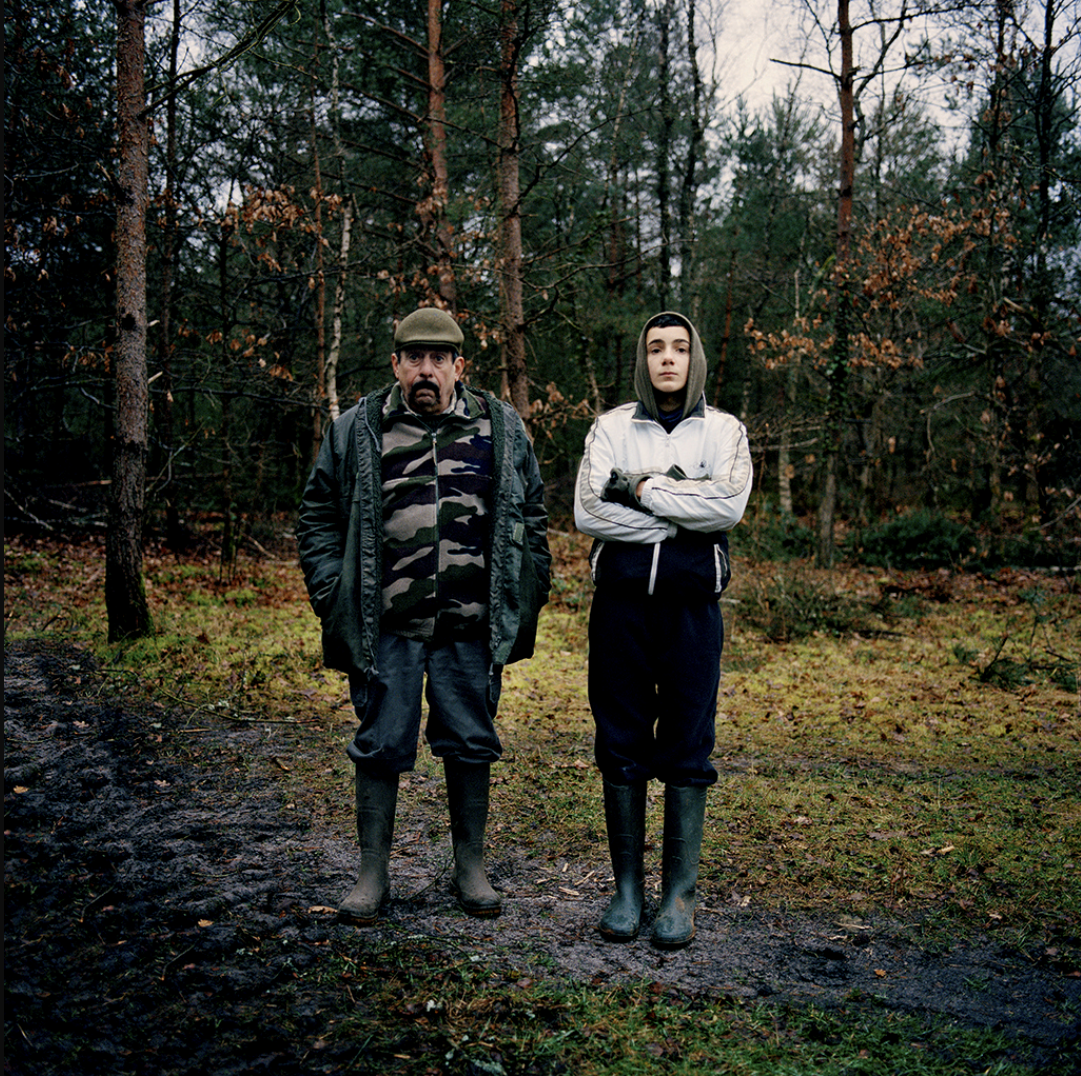 Deux personnes debout dans une forêt, l'une paraît plus âgée avec un chapeau vert et un manteau à motif camouflage, l'autre plus jeune portant une veste blanche avec capuche et des bottes noires, sur un sol en terre humide.