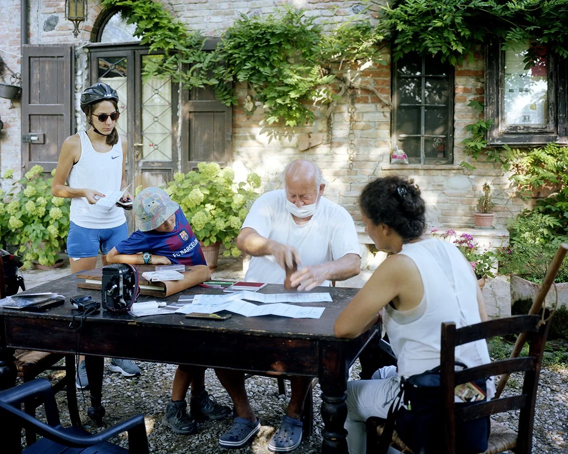A group of five people, including two children, around an outdoor table in front of a stone and green brick house with climbing plants. One of the adults appears to be doing a manual activity, while the others look on or hold their hands.