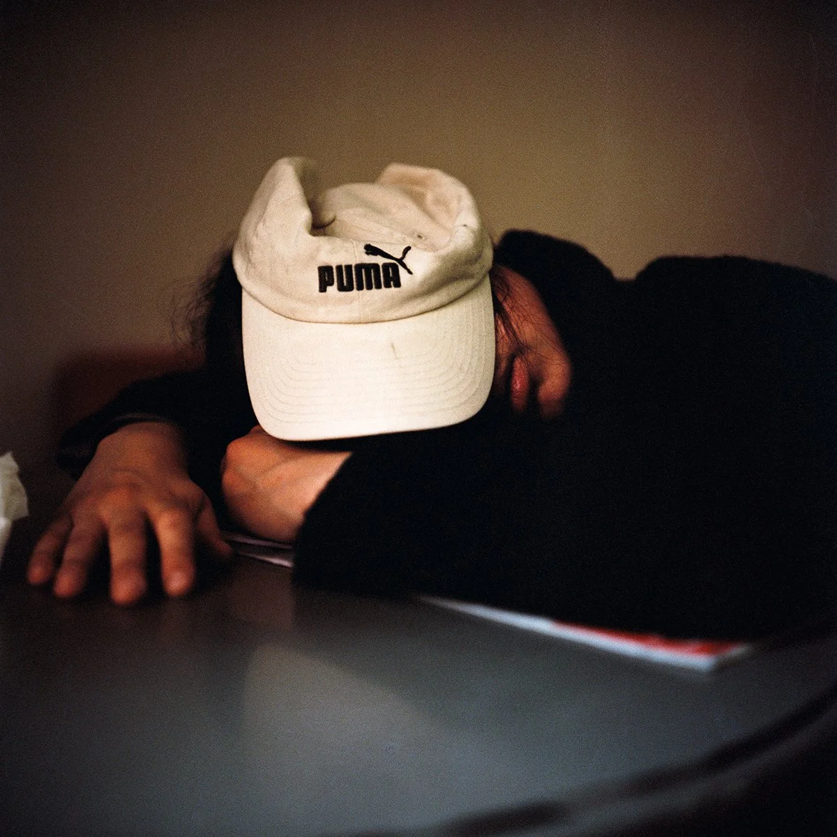 Une personne repose sa tête sur une table, portant une casquette blanche Puma, avec une main posée à plat sur la surface.