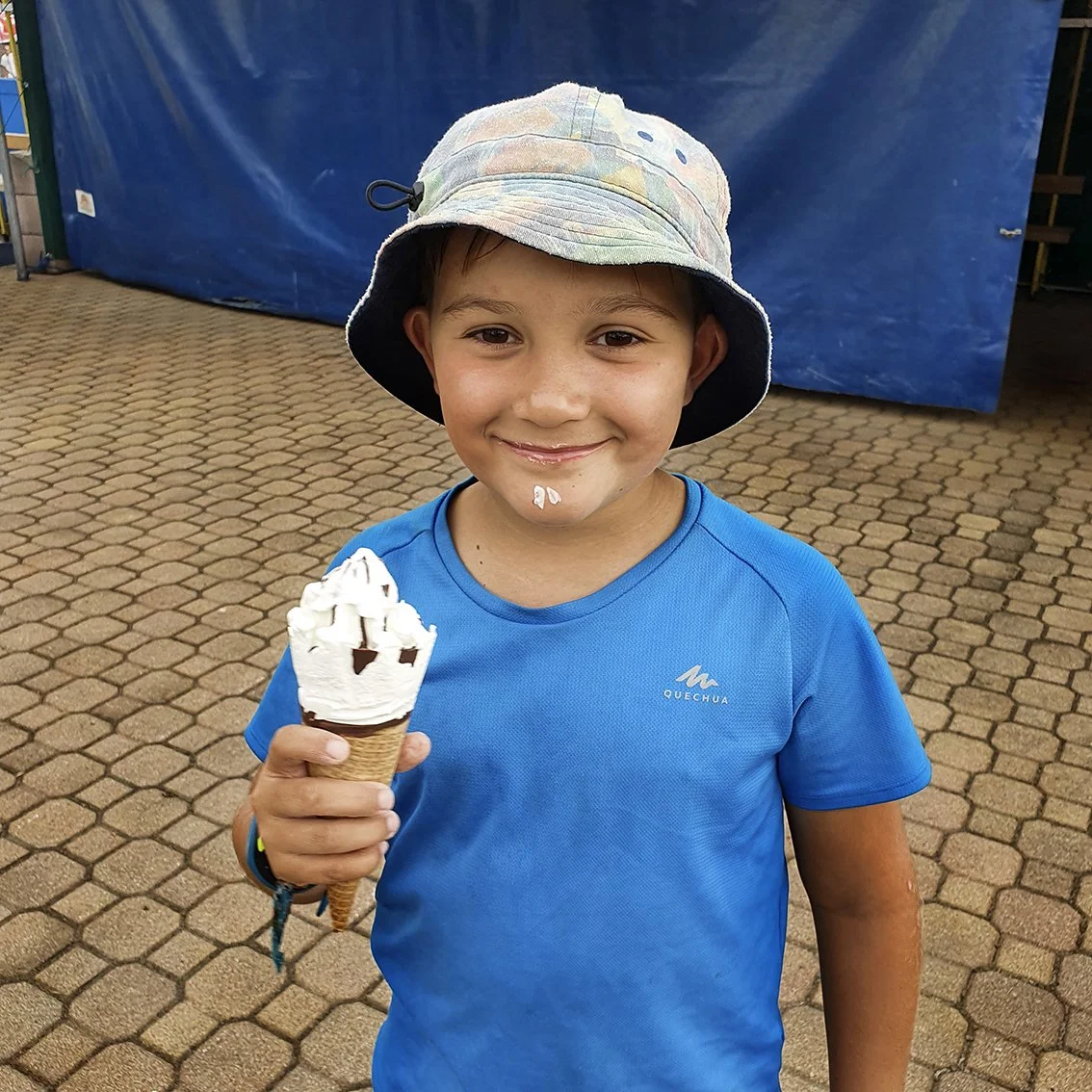 A smiling boy wearing a colorful hat, holding a vanilla ice cream, standing on a paved surface, with a blue background.