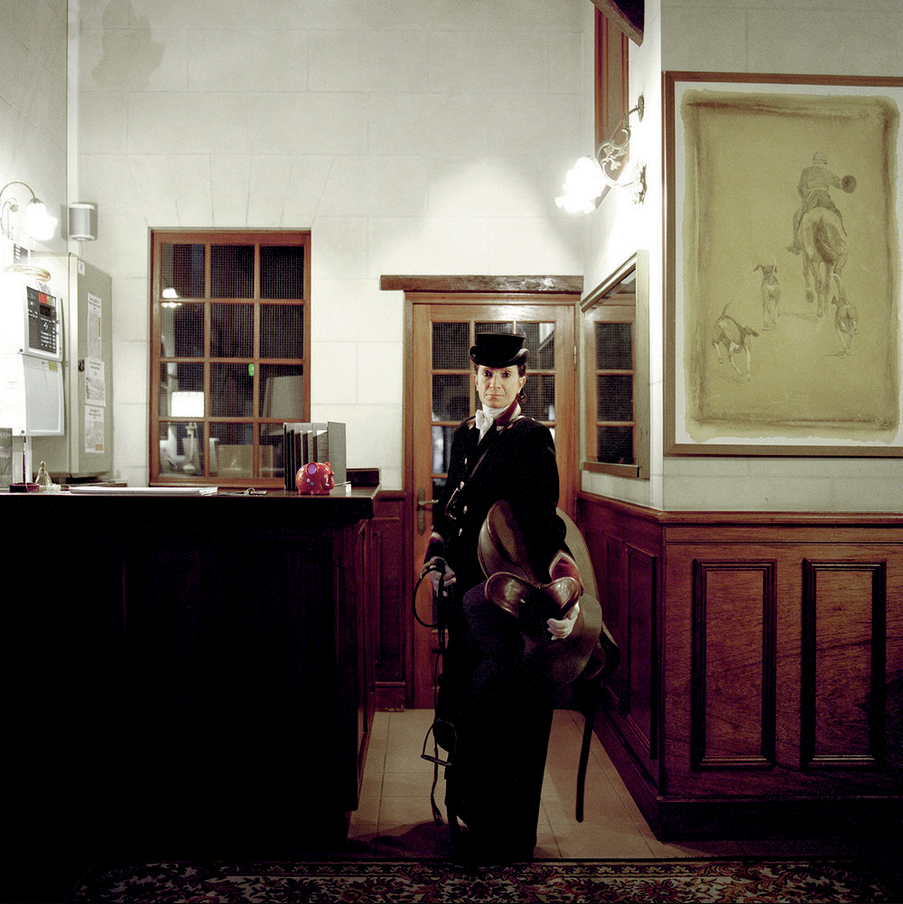 A woman dressed in an old-fashioned uniform, wearing a black hat, stands in an interior with stone walls and wooden panelling, holding a singular batch of leather hats.