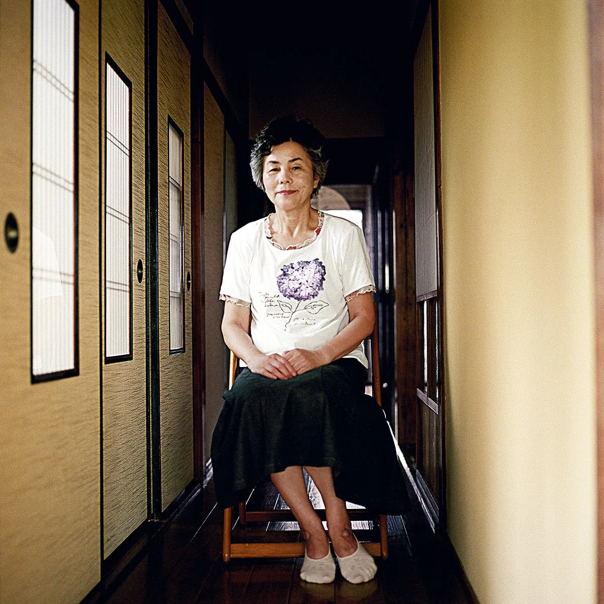 Une femme âgée avec les cheveux courts, assise dans un couloir, portant un T-shirt blanc avec un dessin de fleur violette, une jupe noire, et des chaussettes beige, dans une maison traditionnelle japonaise.