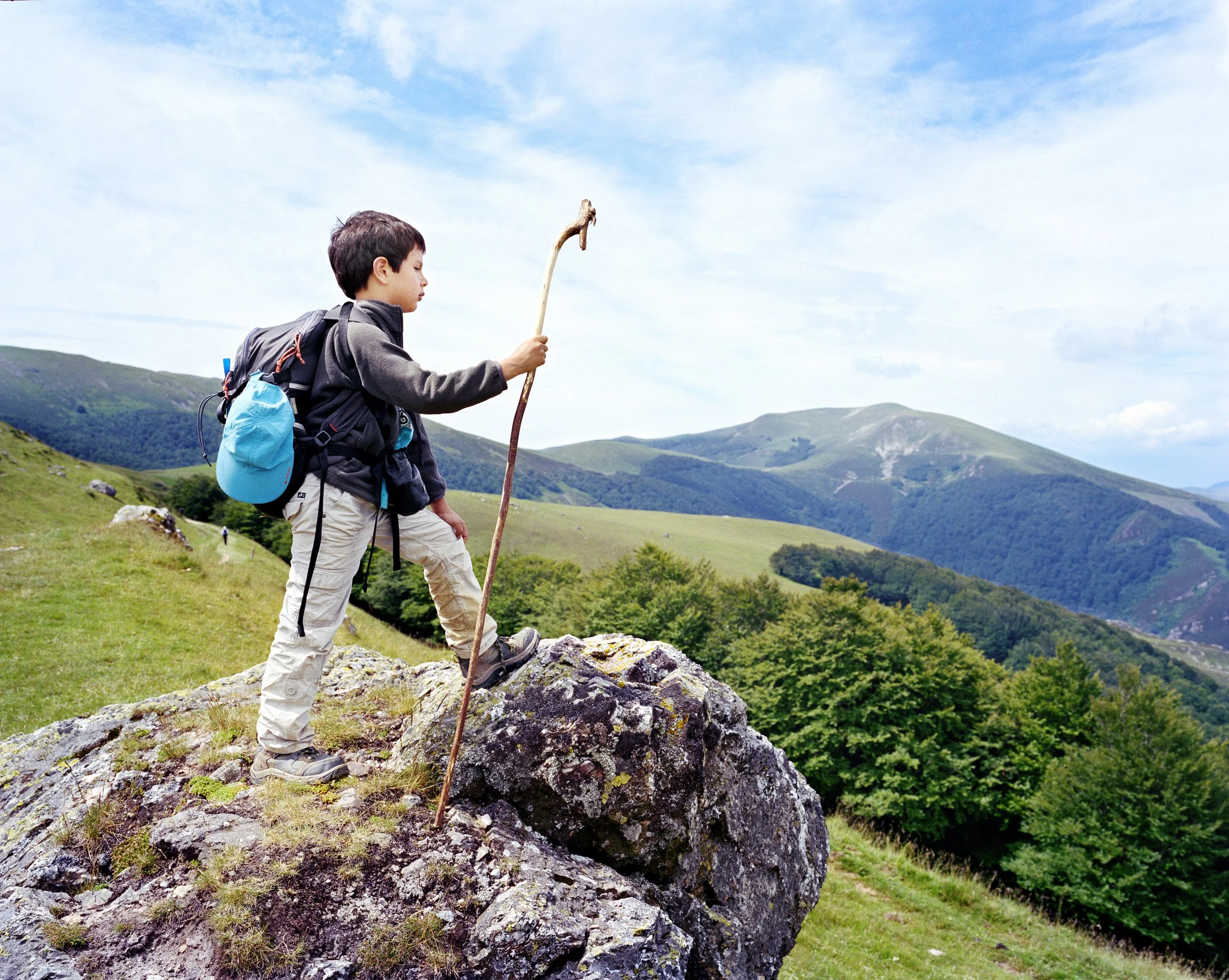 Un garçon avec un sac à dos et une canne sur un rocher en montagne, observant le paysage.