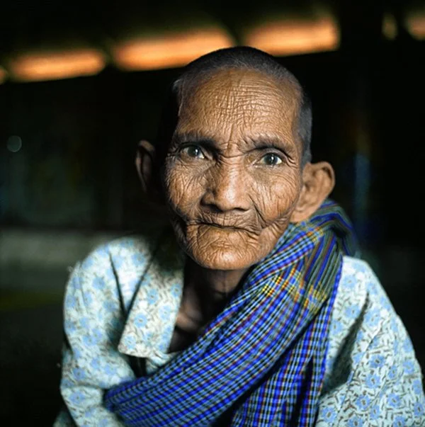 Une vieille femme portant un vêtement coloré, avec un fond sombre, regardant vers la caméra.