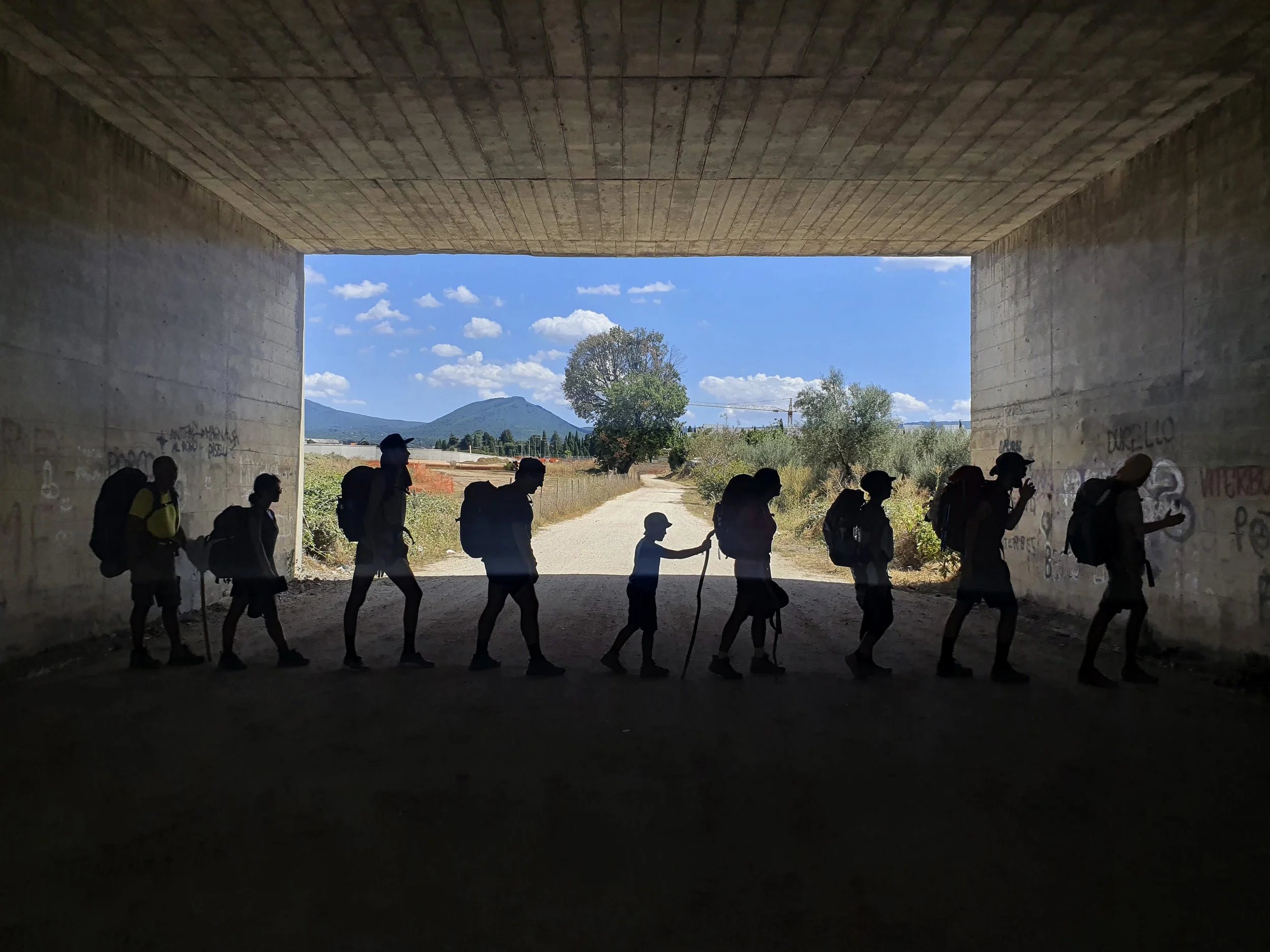 Group of walkers silhouetted under a tunnel, facing daylight, with trees and mountains in the background.