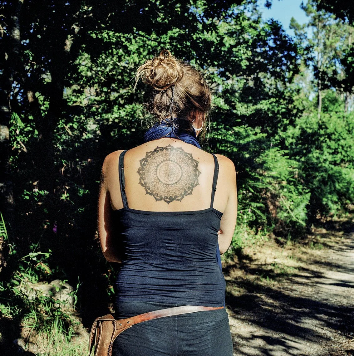 Woman seen from behind wearing a black tank top and mask, with an intricate tattoo on her back, in a natural environment with trees and a dirt road.