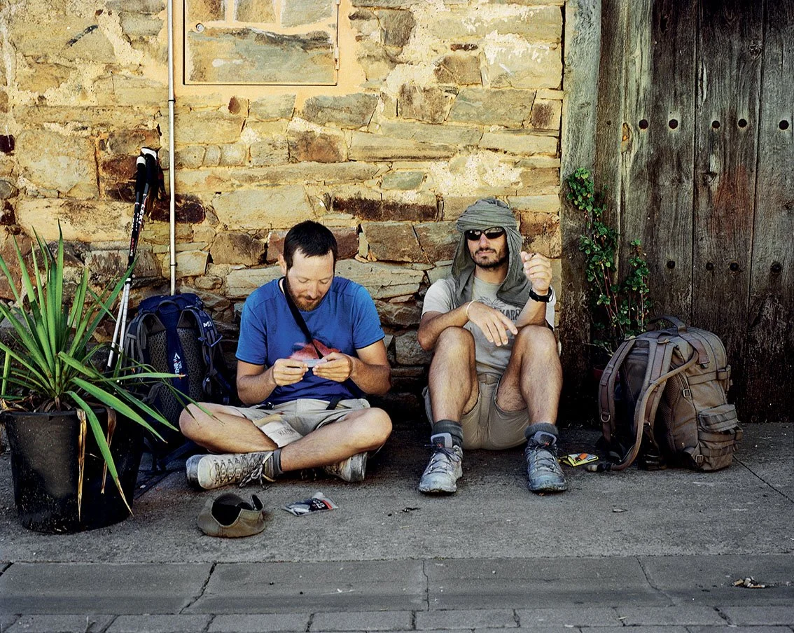 Two hikers sitting against a stone wall, with backpacks, hats and hiking gear, one looking at his phone and the other wearing glasses and a scarf on his head.