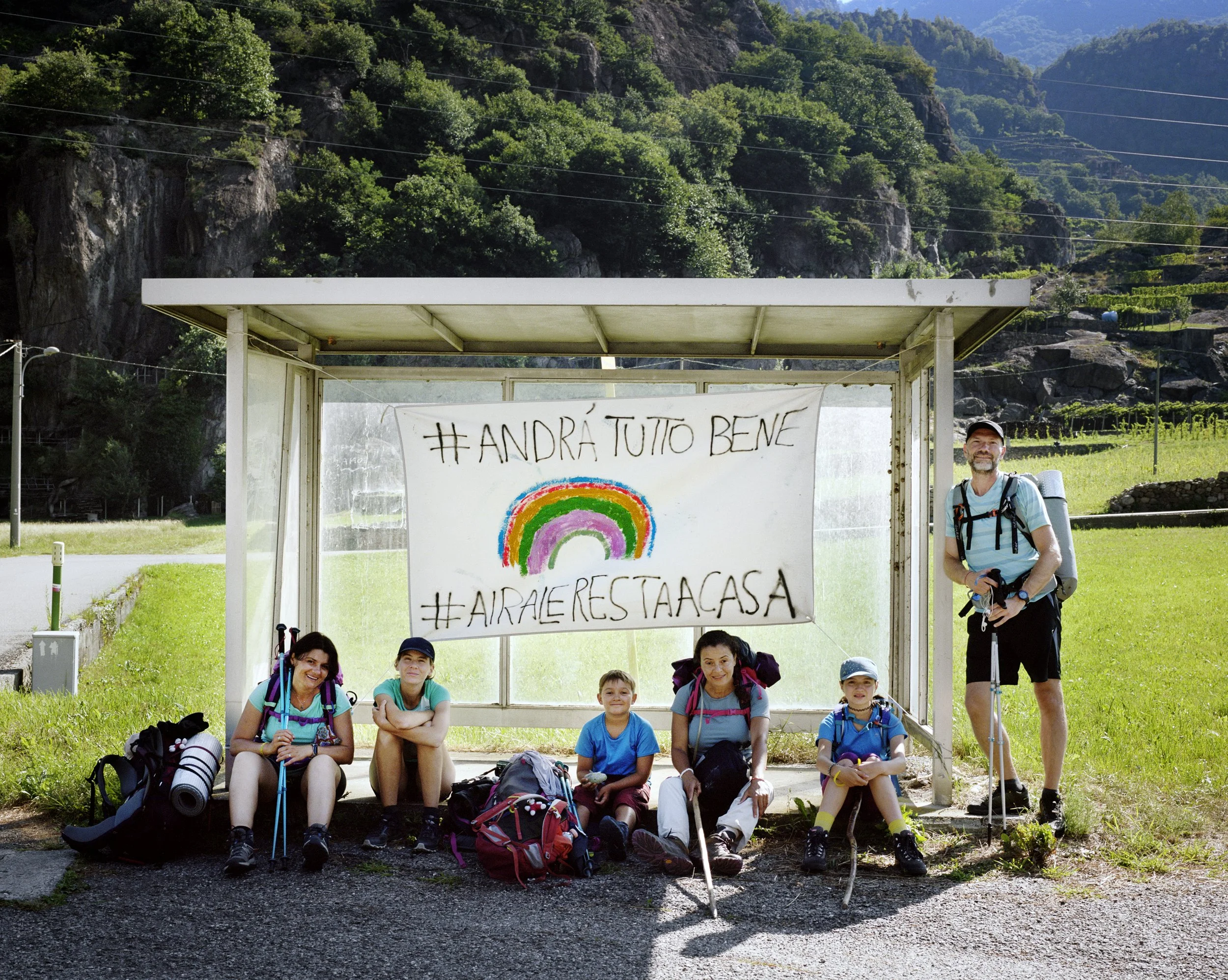 Group of six hiking, sitting or standing in front of a shelter with a flag displaying a rainbow and the hashtags #ANDRÀ TUTTO BENE and #AIRARESTAACASA, in a green mountain landscape.