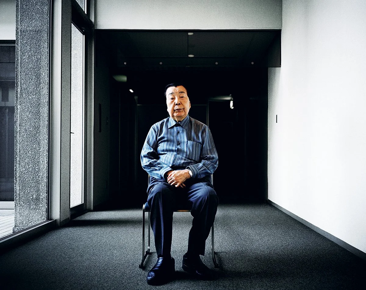 Un homme âgé assis sur une chaise dans un intérieur moderne, lumière naturelle venant d'une fenêtre à gauche, fond sombre derrière lui.
