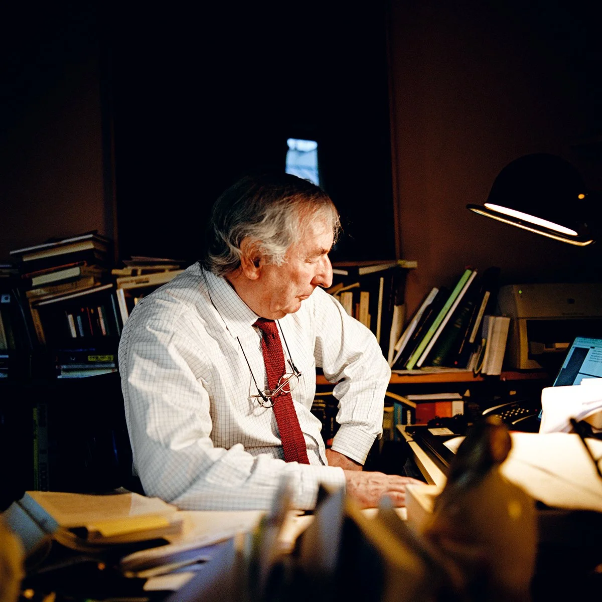 An elderly man sitting at a desk, concentrating on a document or tablet, in an office full of books and papers, lit by a desk lamp.