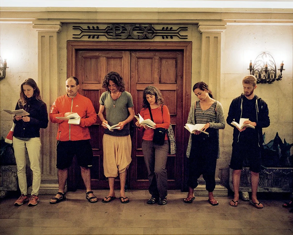 Six people standing, reading books in a room with stone walls and a large wooden door in the background.
