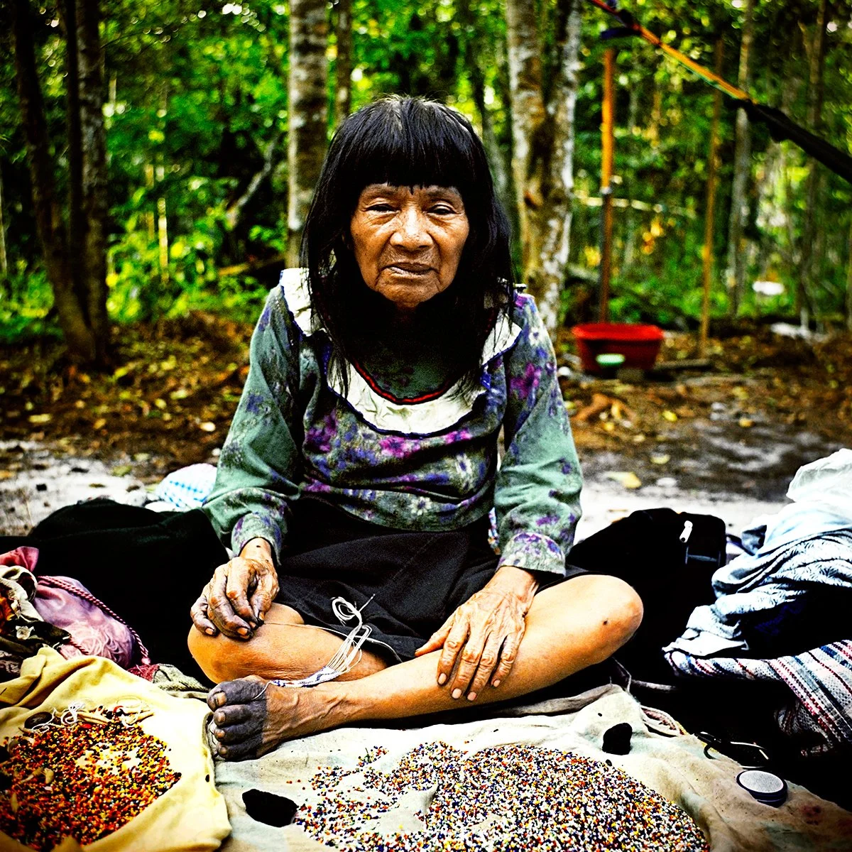 A native woman sits on the ground in a forest, surrounded by colorful beads and bags, with a dense background of green trees.
