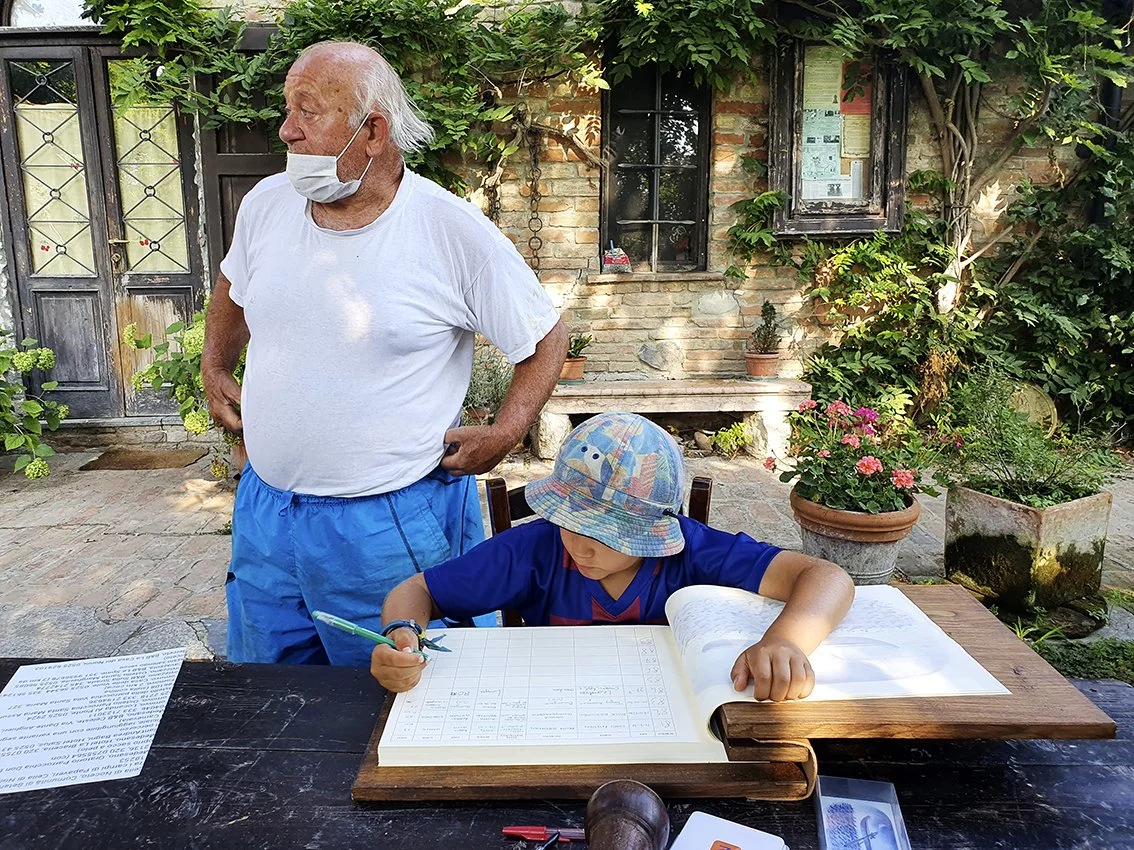 An elderly man wears a face mask and looks to the side, while a young boy writes in an open notebook at an outdoor table, in a garden with plants and a brick house in the background.