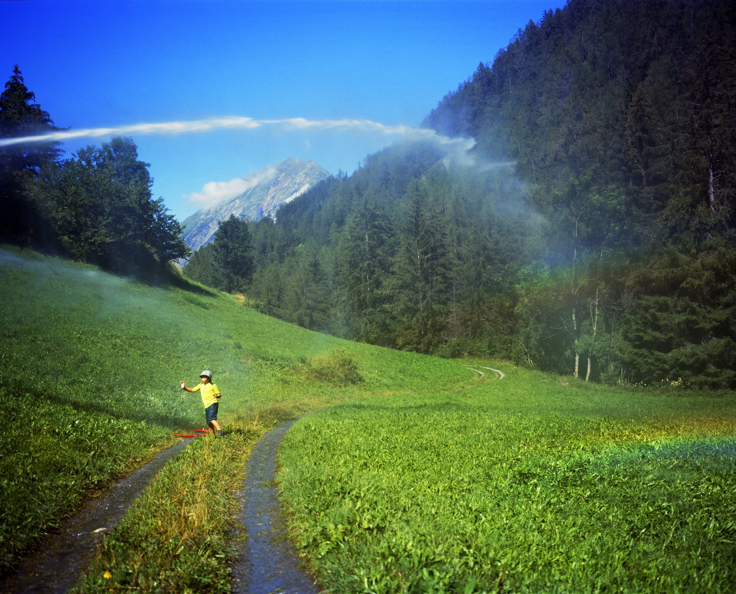 A child plays in a green field surrounded by mountains and forest, with a rainbow visible in the clear blue sky.