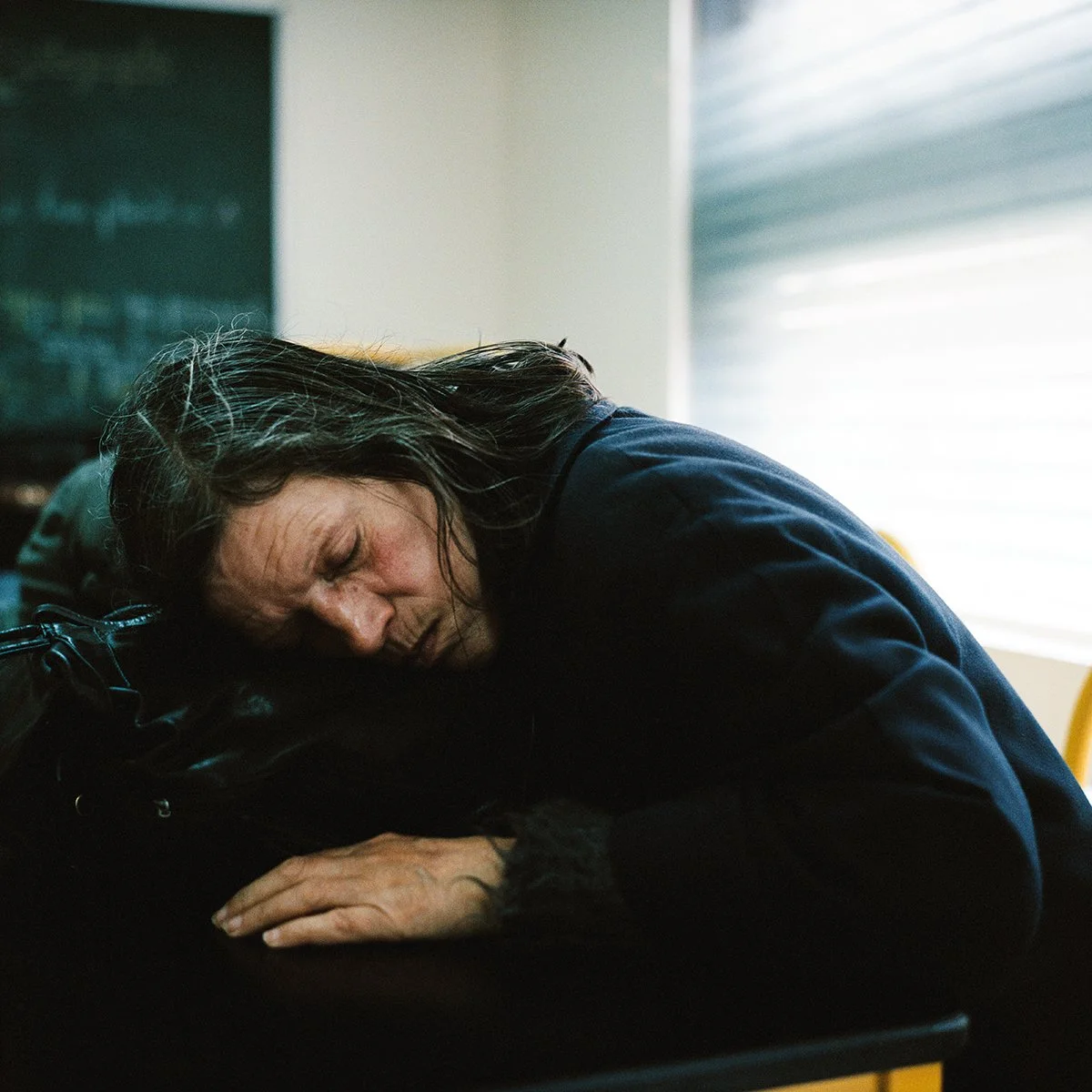 Une femme âgée repose sa tête sur un bureau, semblant dormir ou être épuisée, dans une salle éclairée naturellement par une fenêtre avec un volet roulant.