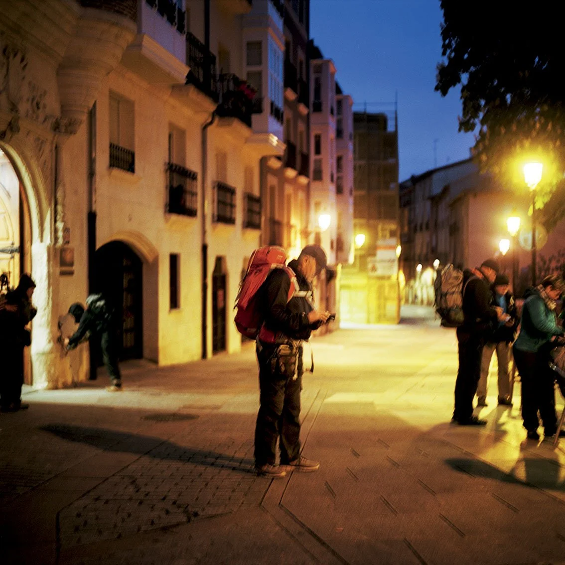 Group of hikers carrying backpacks, meeting on an urban street in the evening, lit by street lamps, with an old building on the left.