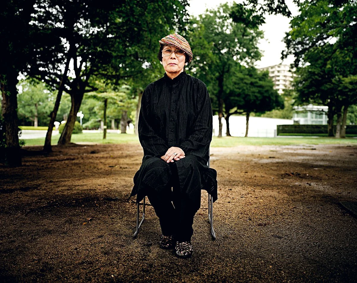 Une femme âgée assise sur une chaise dans un parc verdoyant, portant un chapeau coloré, des lunettes et une tenue noire.